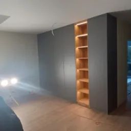 Montaż szafy wnękowej z oświetleniem półek, widoczne narzędzia i lampy budowlane, surowy stan wykończenia wnętrza.