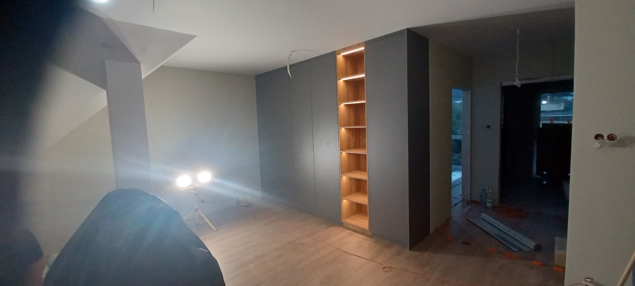 Montaż szafy wnękowej z oświetleniem półek, widoczne narzędzia i lampy budowlane, surowy stan wykończenia wnętrza.