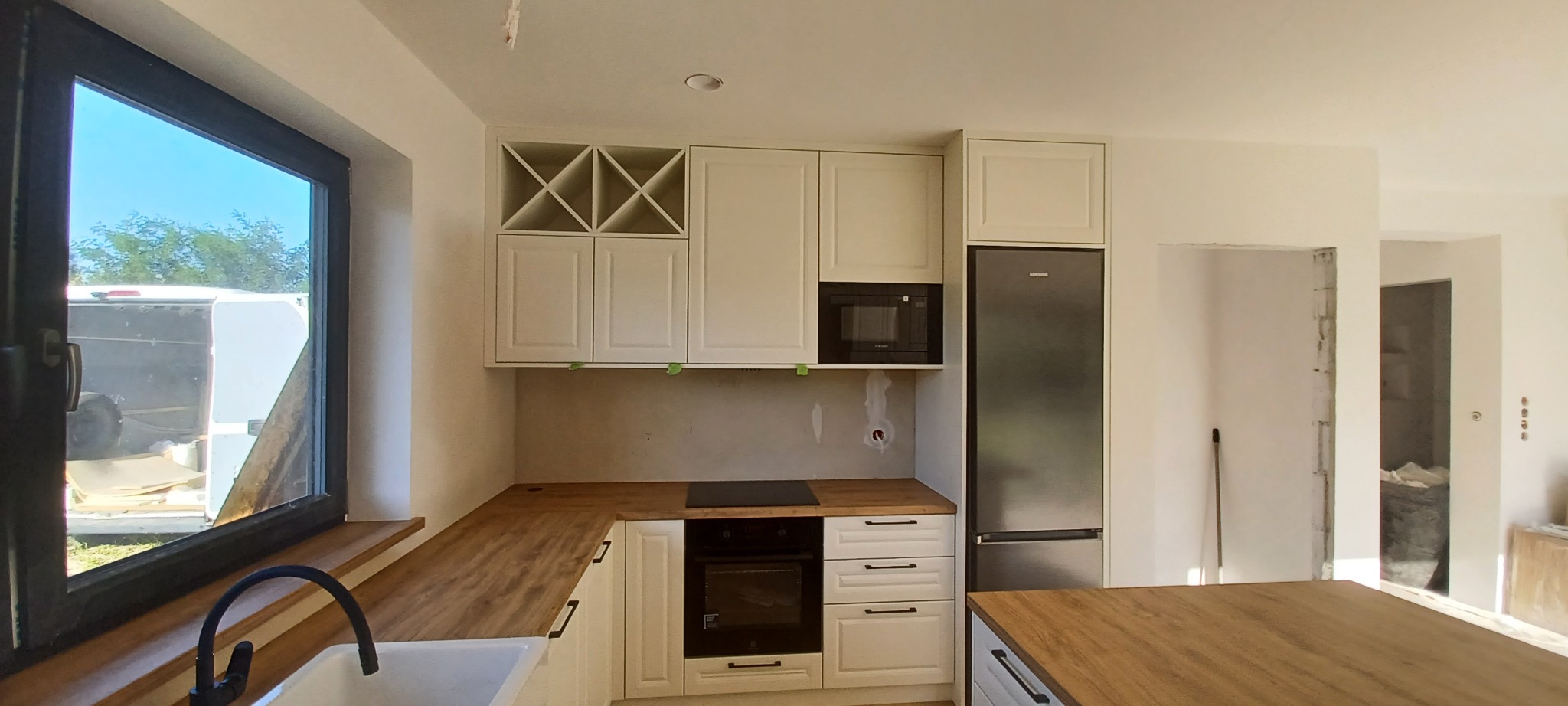 Montaż białej kuchni z drewnianym blatem i wbudowanym sprzętem AGD, widoczne niedokończone ściany i okno z widokiem na dostawczy samochód.