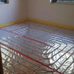 Układanie rur ogrzewania podłogowego na folii izolacyjnej z widocznymi oknami i taśmą malarską wzdłuż ścian.