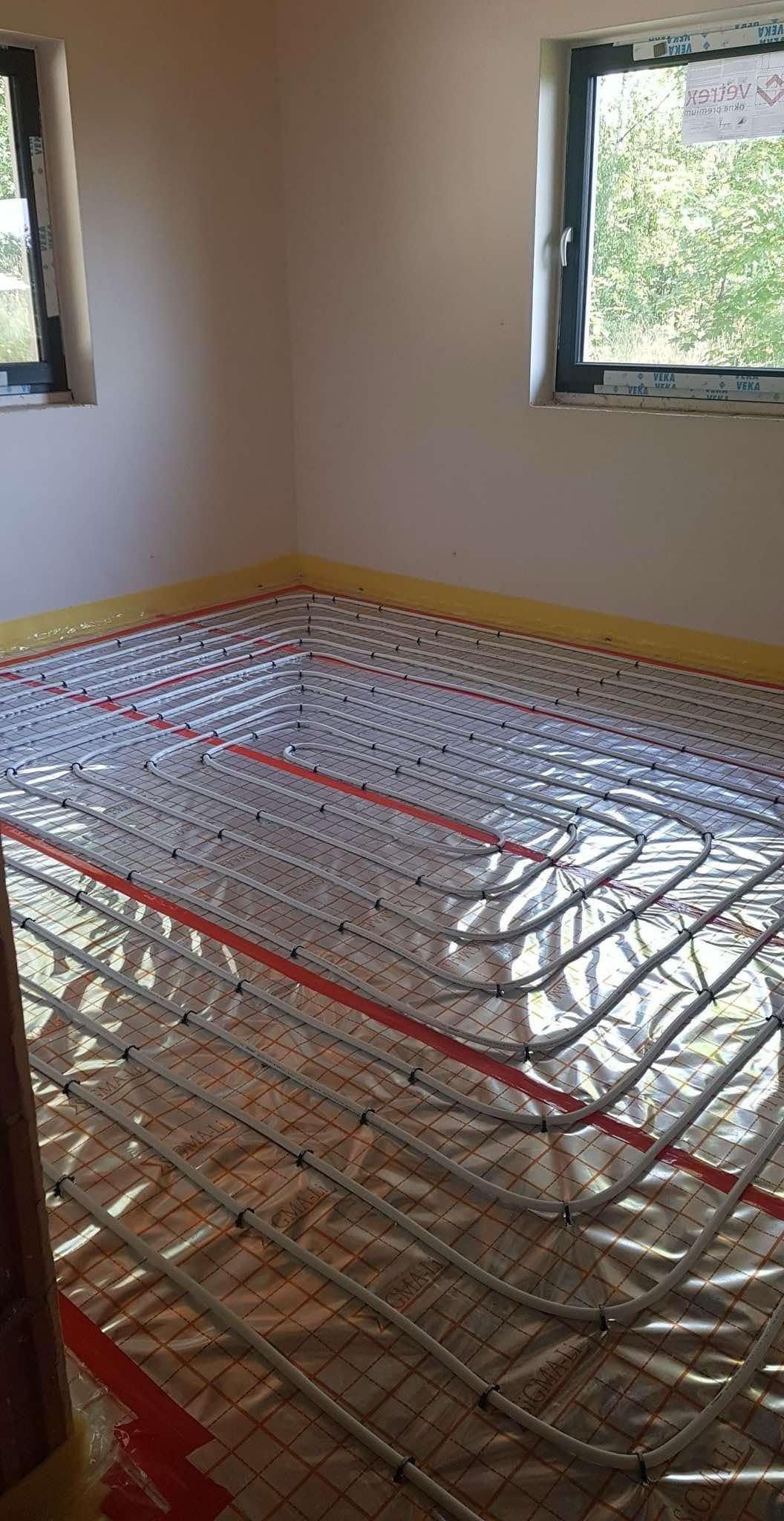 Układanie rur ogrzewania podłogowego na folii izolacyjnej z widocznymi oknami i taśmą malarską wzdłuż ścian.