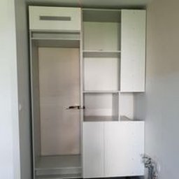 Rafal Sarnowski - Biała, częściowo zmontowana szafa z otwartymi półkami i szufladą w górnej części, widoczne elementy montażowe i przewody na podłodze, surowe ściany w tle.