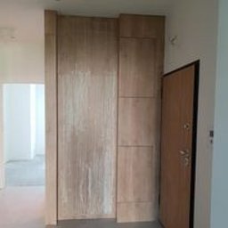 Rafal Sarnowski - Wnętrze w trakcie wykańczania: Duża, pionowa konstrukcja z paneli drewnopodobnych w odcieniach beżu i brązu, prawdopodobnie element zabudowy wnęki, sąsiaduje z otwartymi drzwiami wejściowymi.