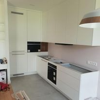 Jasna, minimalistyczna kuchnia z białymi frontami szafek, szarym blatem i sprzętem AGD w zabudowie. Widoczne elementy wykończenia wnętrza, surowy beton na podłodze i różowa ściana w tle.