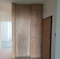 Wnętrze w trakcie wykańczania: Duża, pionowa konstrukcja z paneli drewnopodobnych w odcieniach beżu i brązu, prawdopodobnie element zabudowy wnęki, sąsiaduje z otwartymi drzwiami wejściowymi.