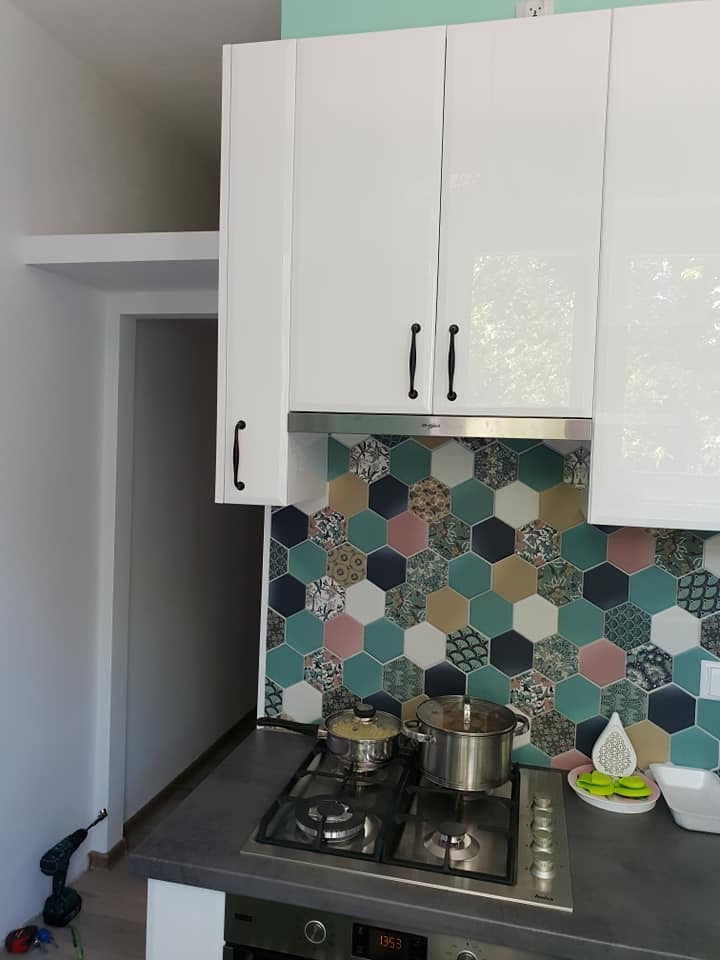 Białe, lakierowane szafki kuchenne z czarnymi uchwytami nad kuchenką gazową z garnkami, tło stanowi mozaika z heksagonalnych płytek w odcieniach zieleni, różu i brązu.