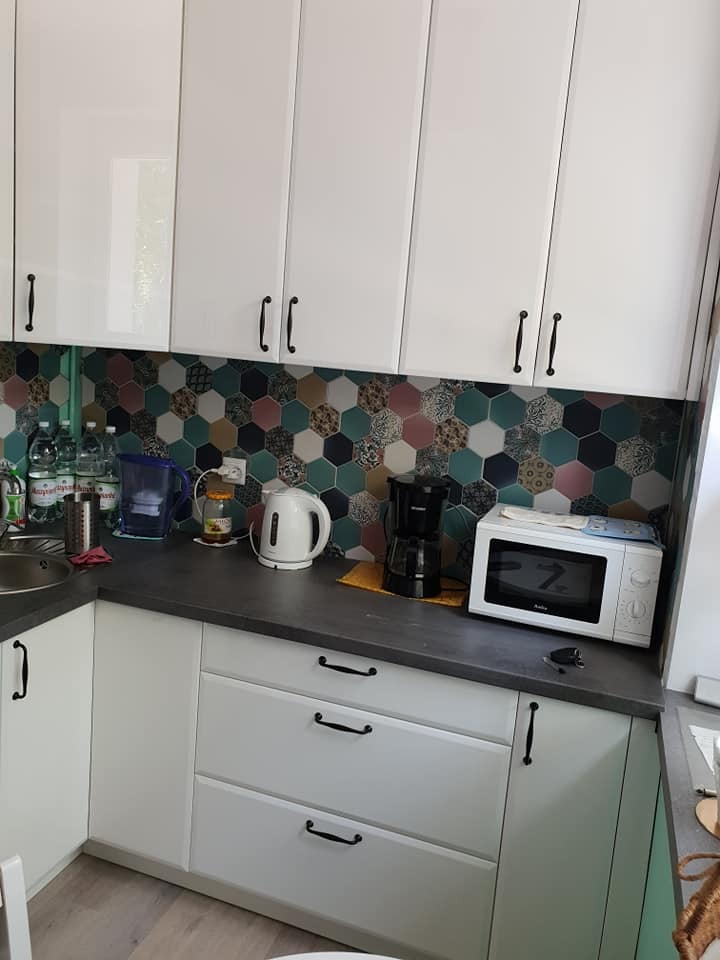 Jasna kuchnia z białymi szafkami, ciemnym blatem i mozaiką heksagonalną na ścianie. Widoczne sprzęty AGD: czajnik, ekspres do kawy, mikrofalówka.