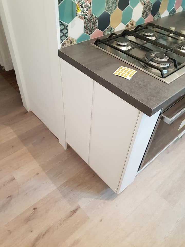 Fragment zabudowy kuchennej z białymi frontami, szarym blatem i płytą gazową. Na blacie naklejka z żółtymi kropkami. W tle heksagonalne płytki w odcieniach turkusu, granatu i różu.