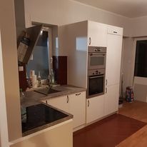 Biała zabudowa kuchenna z piekarnikiem i mikrofalówką w nowoczesnym stylu, widok z boku na blat z płytą indukcyjną i okap, brązowe akcenty na ścianie i podłodze.