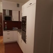 Biała zabudowa kuchenna z ciemnoczerwonym panelem nad blatem, widoczna kuchenka mikrofalowa i okap, ujęcie z boku ukazujące część szafek i sprzętów AGD.