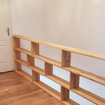 Surowy drewniany regał na wymiar w trakcie montażu, widoczne niedoskonałości obróbki drewna, jasne wnętrze z parkietem i białą ścianą, brak wykończenia.