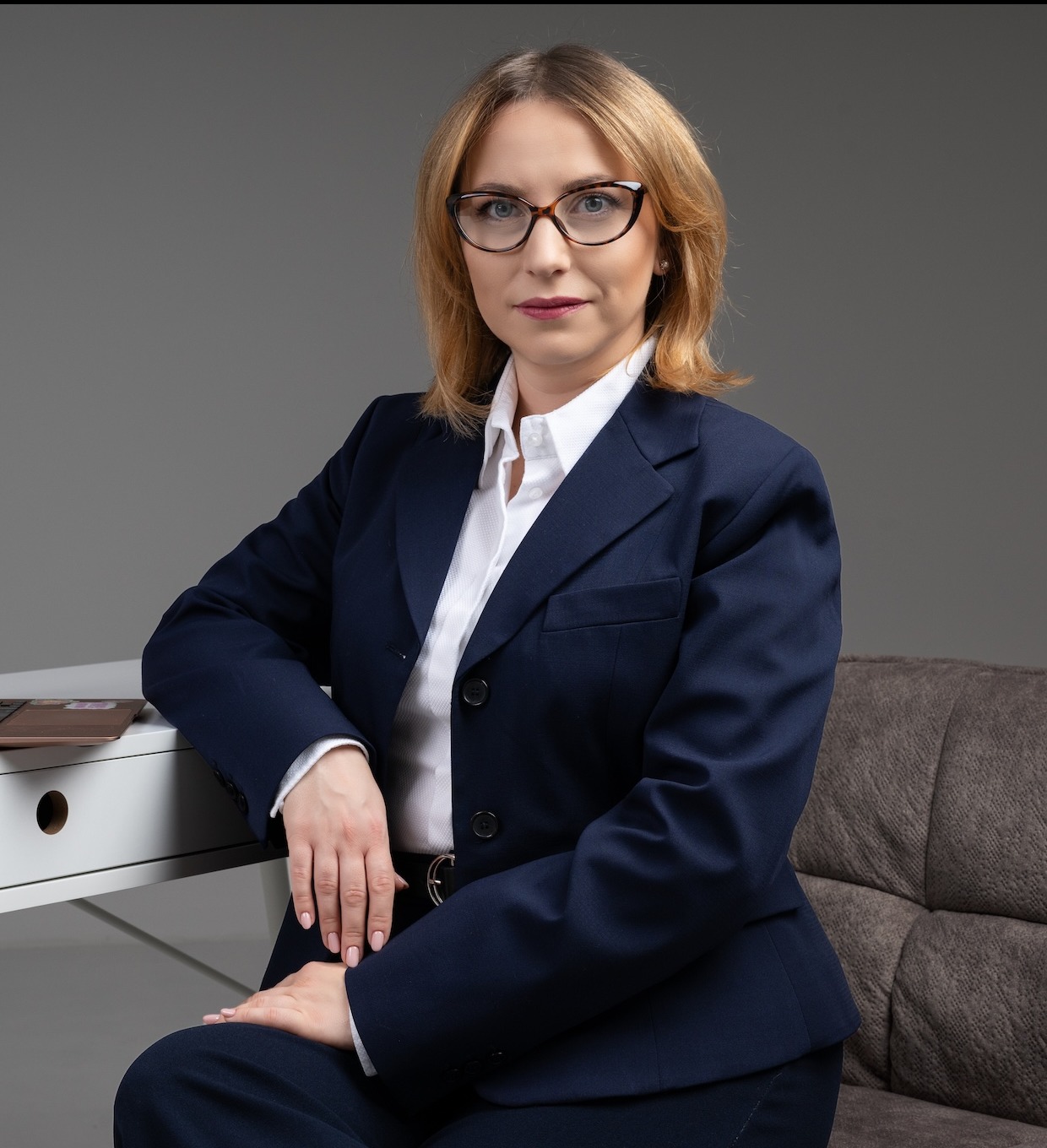 Portret kobiety w okularach i granatowym garniturze, siedzącej przy białym biurku z laptopem, na szarym tle.