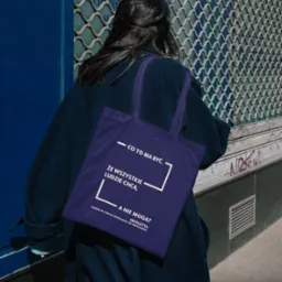 Osoba z ciemnymi włosami, ubrana w granatowy płaszcz, niesie fioletową torbę z nadrukowanym tekstem i logo fundacji, na tle zamkniętej, metalowej rolety z niebieskimi elementami.