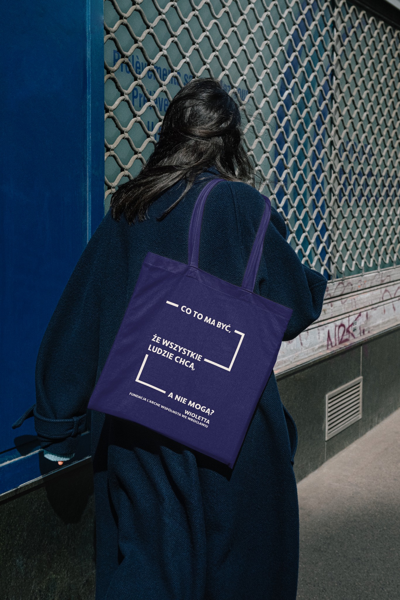 Osoba z ciemnymi włosami, ubrana w granatowy płaszcz, niesie fioletową torbę z nadrukowanym tekstem i logo fundacji, na tle zamkniętej, metalowej rolety z niebieskimi elementami.