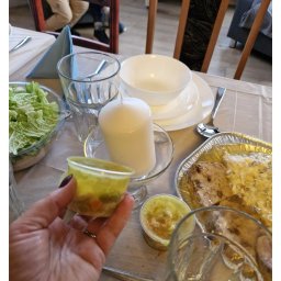 Katarzyna Wittbrodt - Stół zastawiony naczyniami i jedzeniem, w dłoni przezroczysty pojemnik z sałatką warzywną, widoczne danie mięsne w folii aluminiowej, sałatka w szklanej misce, biała świeca i szklanki.