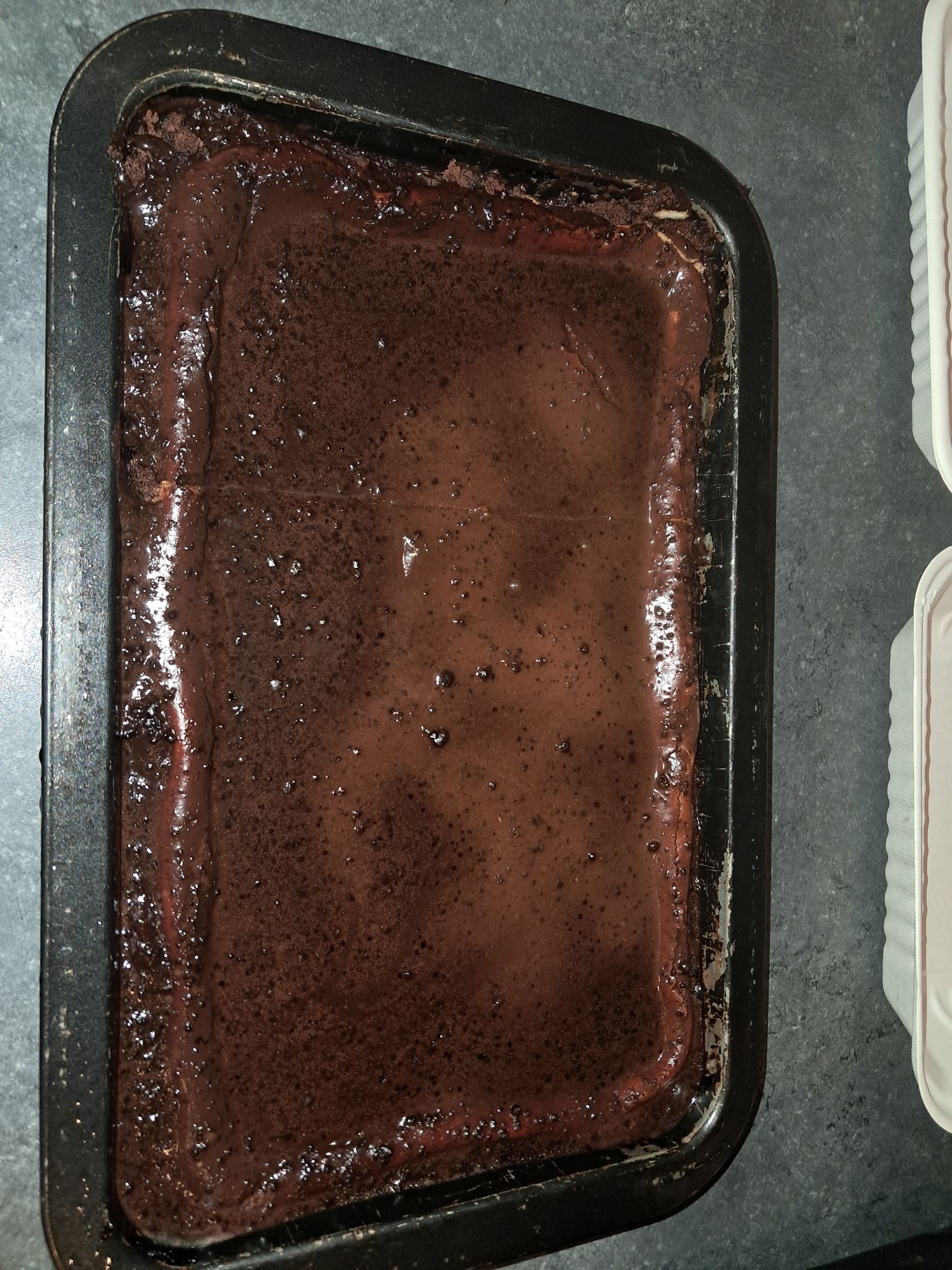 Ciemne, czekoladowe ciasto w prostokątnej, czarnej formie do pieczenia, z widoczną nierówną powierzchnią i błyszczącą polewą, obok pojemniki na wynos.