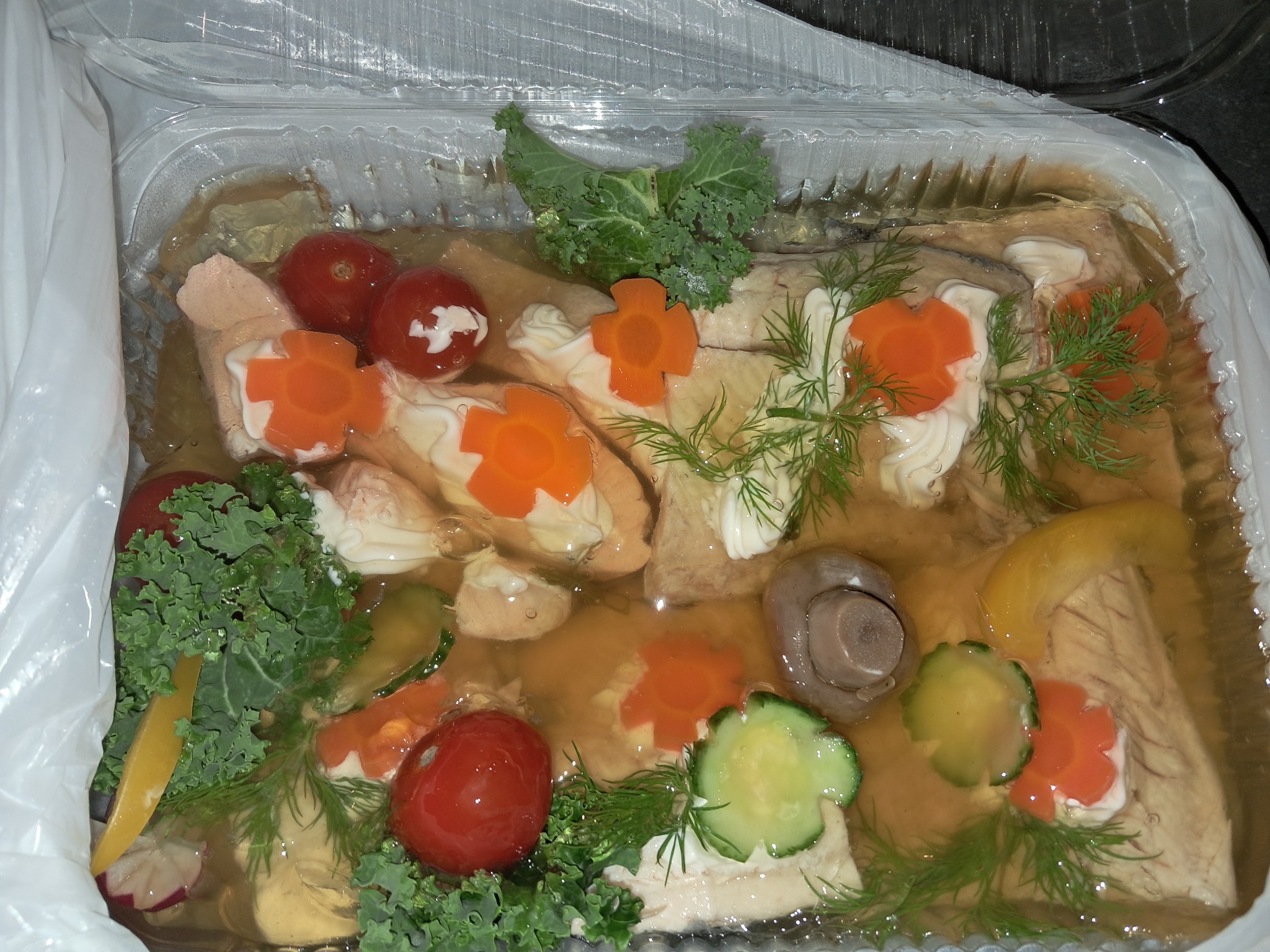 Ryba w galarecie, udekorowana marchewką w kształcie kwiatów, pomidorkami koktajlowymi, ogórkiem, grzybami, koperkiem i jarmużem, zapakowana w plastikowe pudełko.