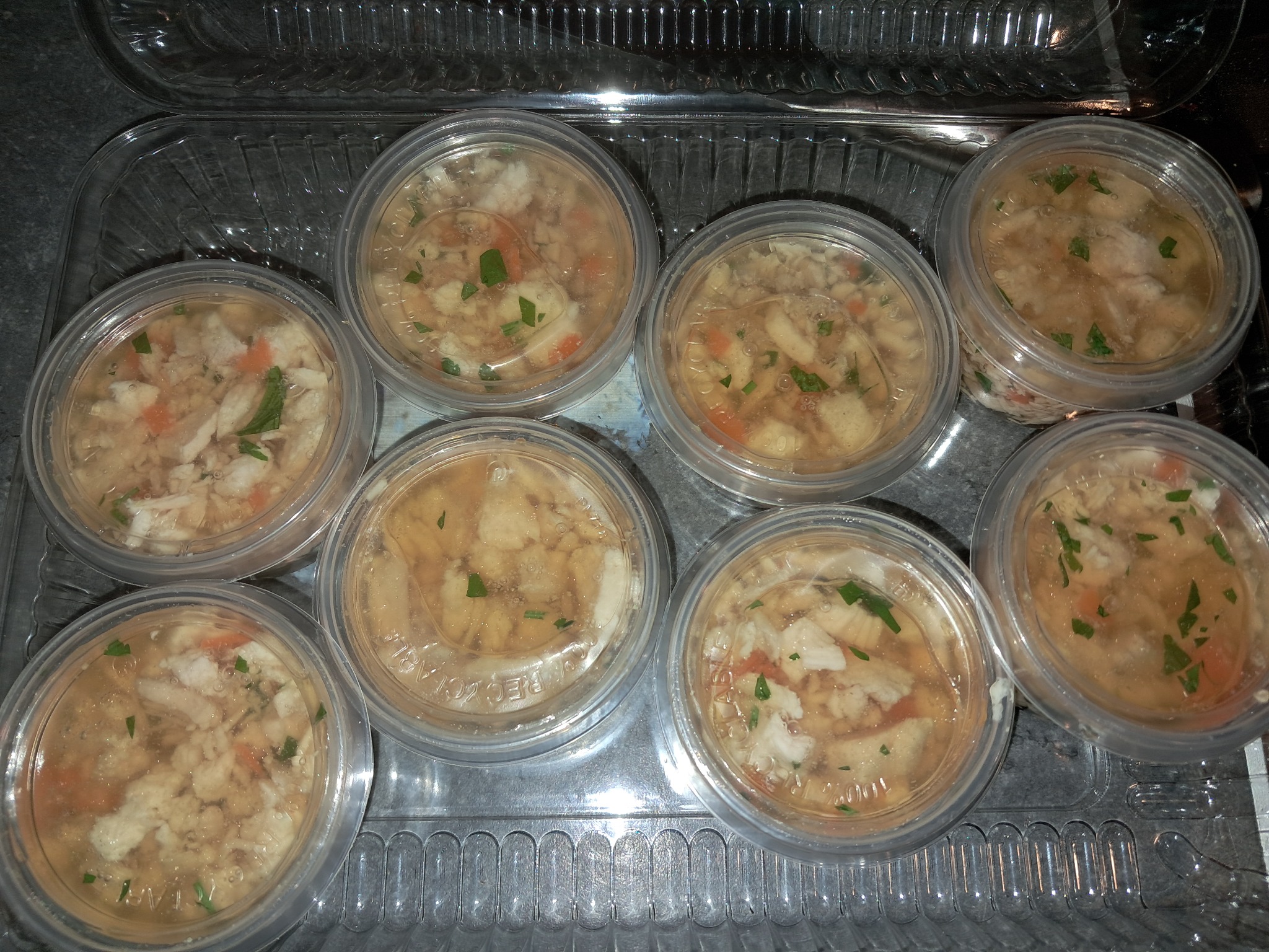 Galaretka z kurczaka w przezroczystych pojemnikach, posypana zieloną pietruszką, ułożona w plastikowym opakowaniu zbiorczym.