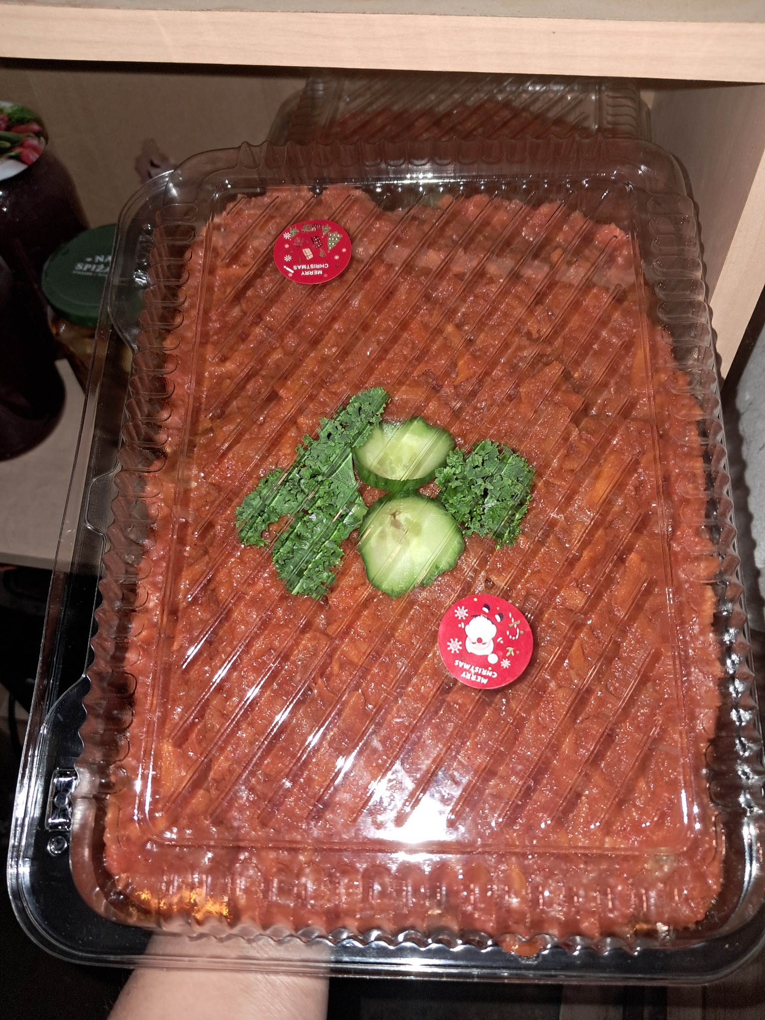 Sałatka warzywna w przezroczystym plastikowym pojemniku, udekorowana plasterkami ogórka i listkami zielonej pietruszki, z naklejkami 'Merry Christmas' na wierzchu, umieszczona na drewnianej półce.