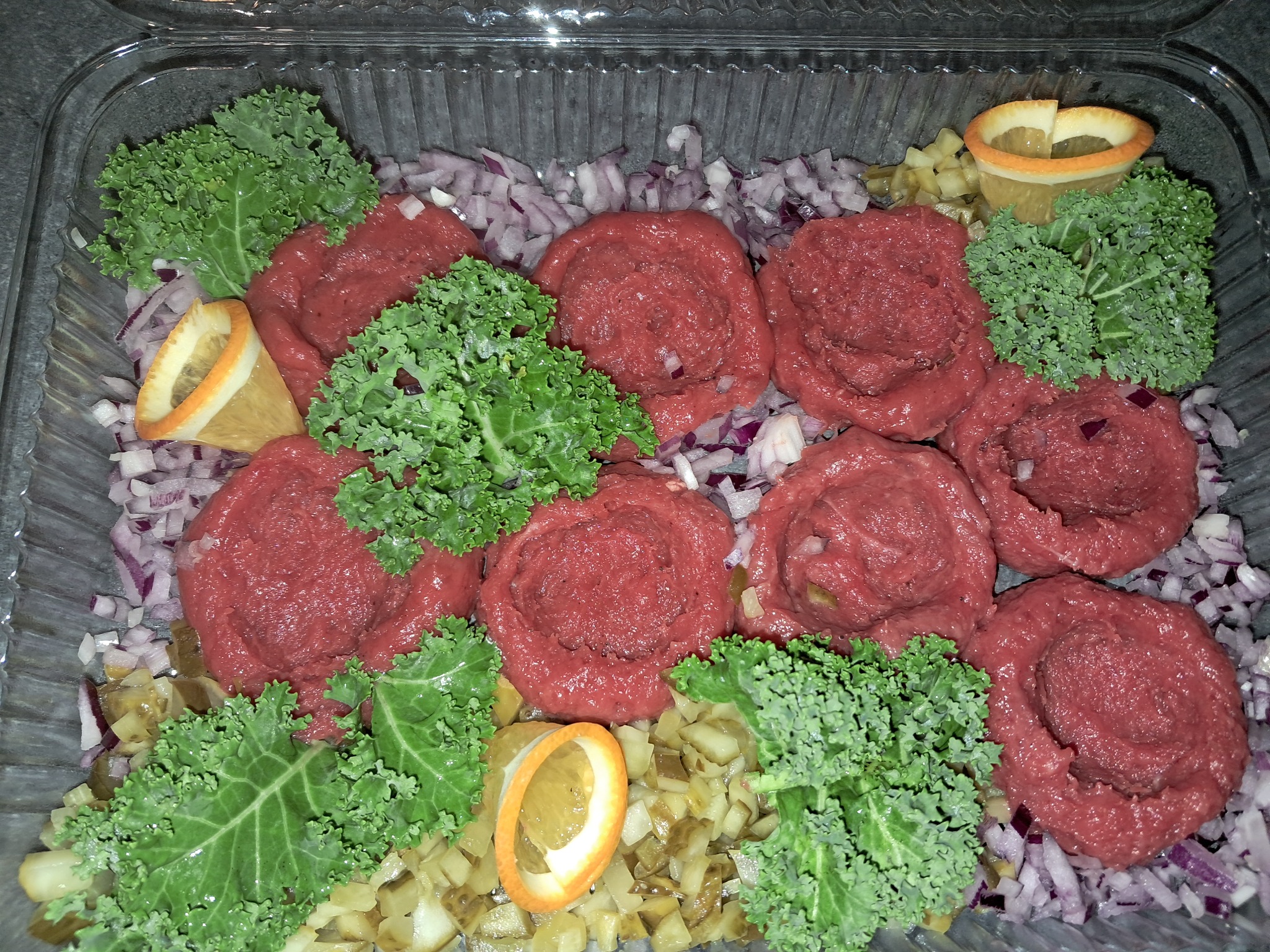 Tatarskie steki uformowane w róże, udekorowane liśćmi jarmużu i plasterkami pomarańczy, ułożone na posiekanej cebuli i ogórkach w przezroczystym pojemniku.