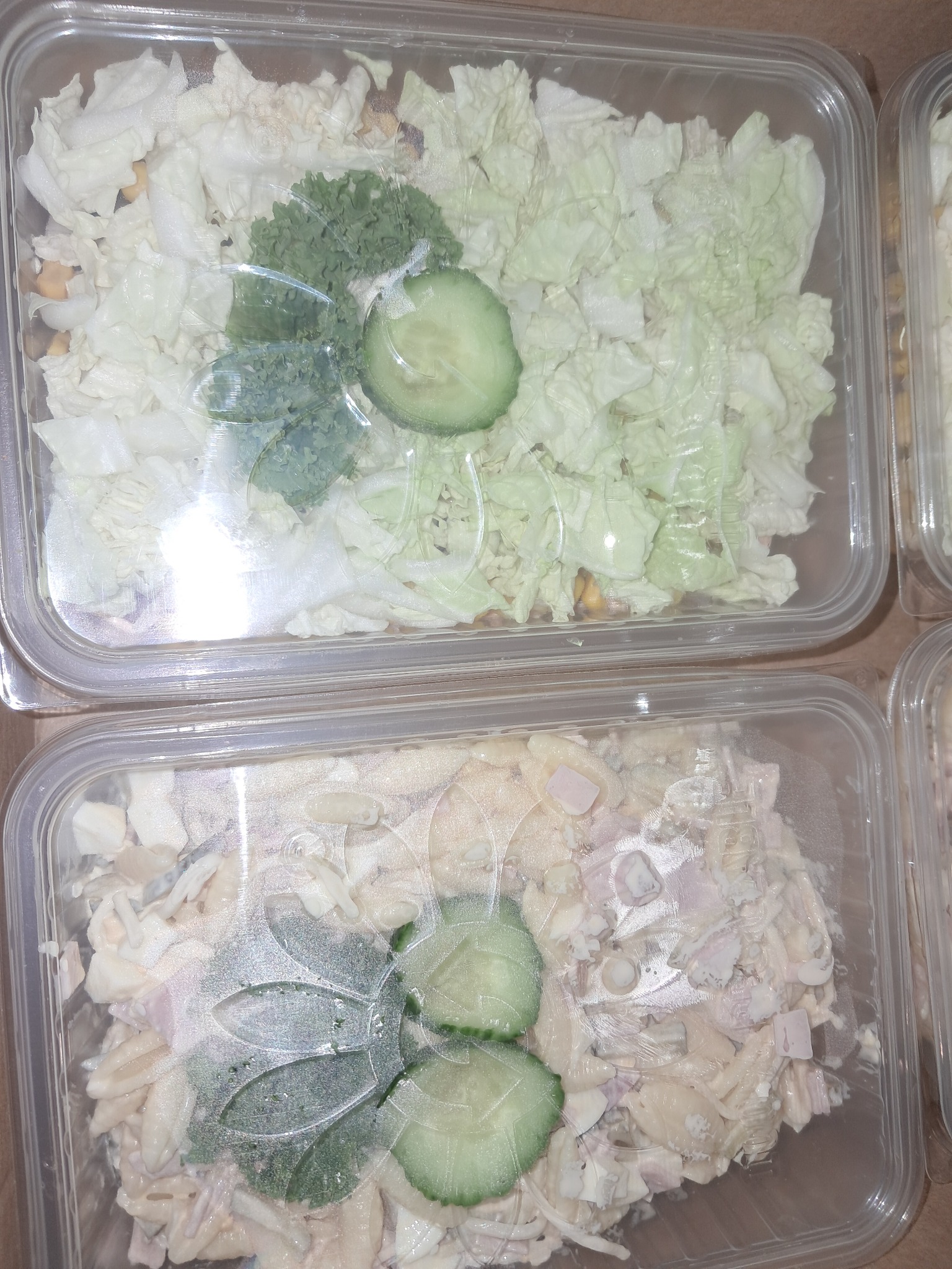 Dwa plastikowe pojemniki z sałatkami pod folią: jeden z sałatą pekińską i zielonymi warzywami, drugi z sałatką makaronową z majonezem i dodatkami. Oba pojemniki udekorowane plasterkami ogórka.