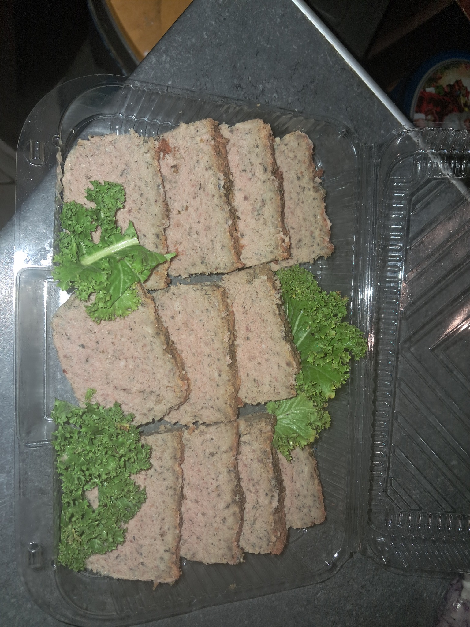 Krojony pasztet w przezroczystym plastikowym pojemniku, udekorowany kędzierzawą sałatą, leżący na szarym blacie.