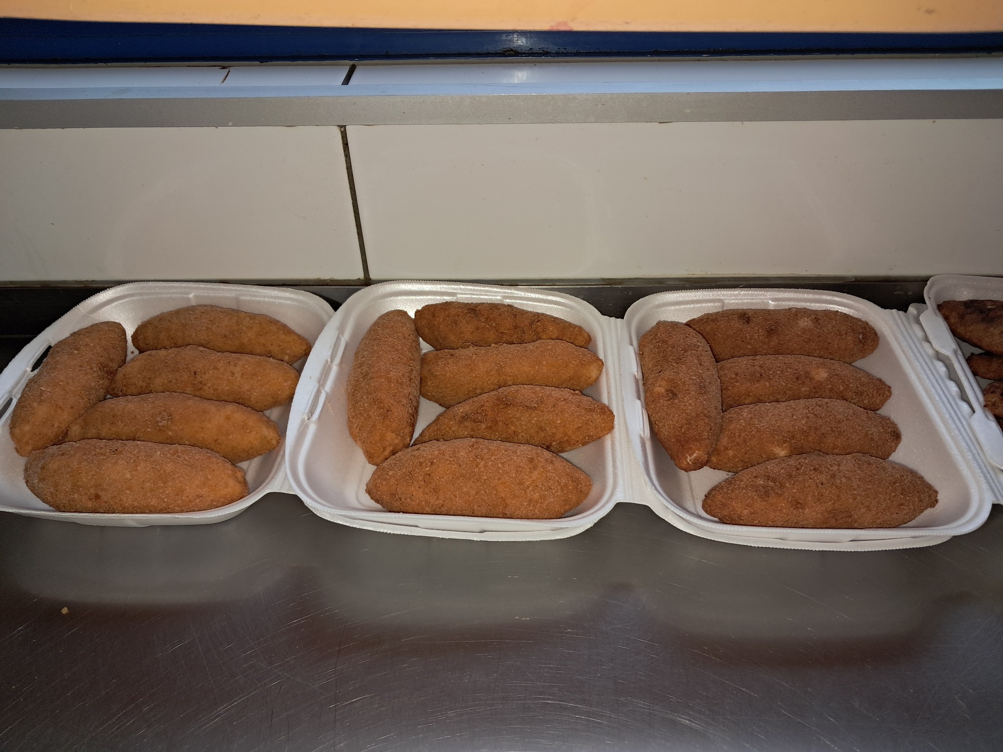 Pączki w podłużnym kształcie, posypane brązowym cukrem, ułożone w trzech białych, styropianowych pojemnikach na metalowym blacie.