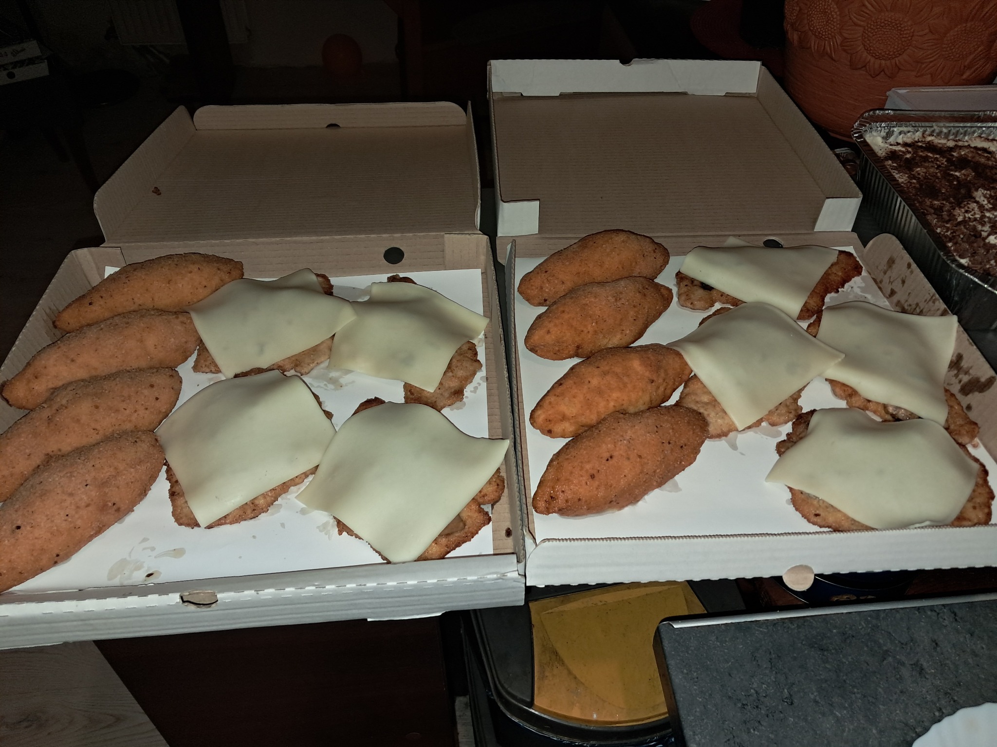 Dwa otwarte kartony po pizzy zawierające kotlety schabowe z serem, obok blaszka z deserem tiramisu, prawdopodobnie dostarczone na imprezę.