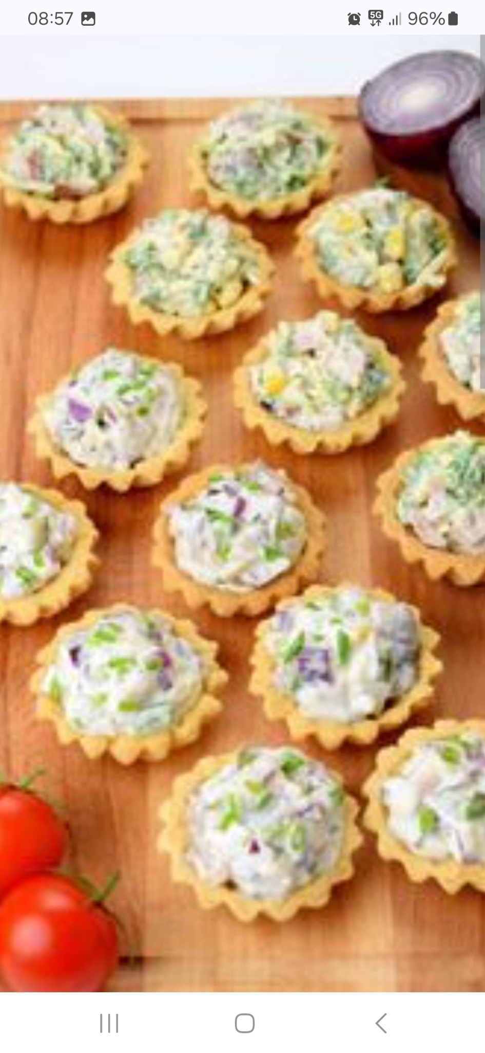 Mini tartaletki z różnymi nadzieniami (sałatka warzywna, pasta jajeczna) udekorowane szczypiorkiem, ułożone na drewnianej desce, z pomidorami i przekrojoną czerwoną cebulą w tle.