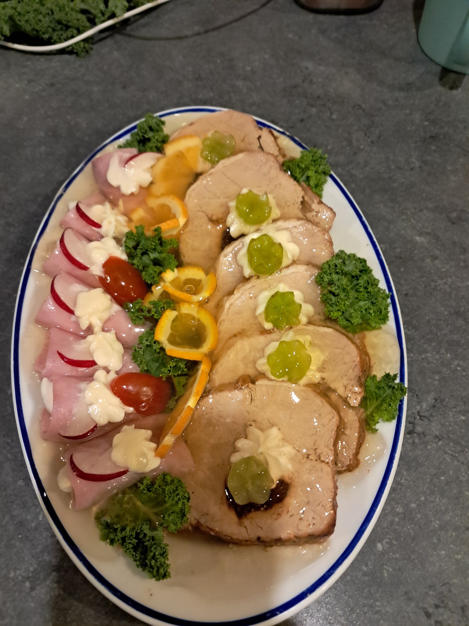 Elegancka kompozycja wędlin, roladek z szynki i pieczonego mięsa na białym półmisku z dekoracją z pomarańczy, rzodkiewek, pomidorków i zielonego jarmużu.