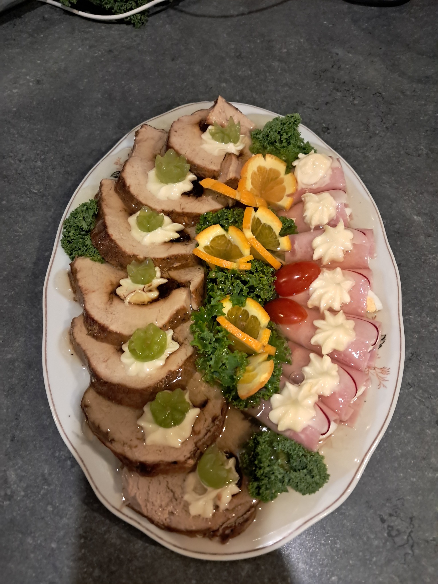 Elegancka, owalna patera z pieczonym mięsem w plastrach, roladkami z szynki, pomidorkami koktajlowymi i dekoracjami z pomarańczy oraz zielonego jarmużu.