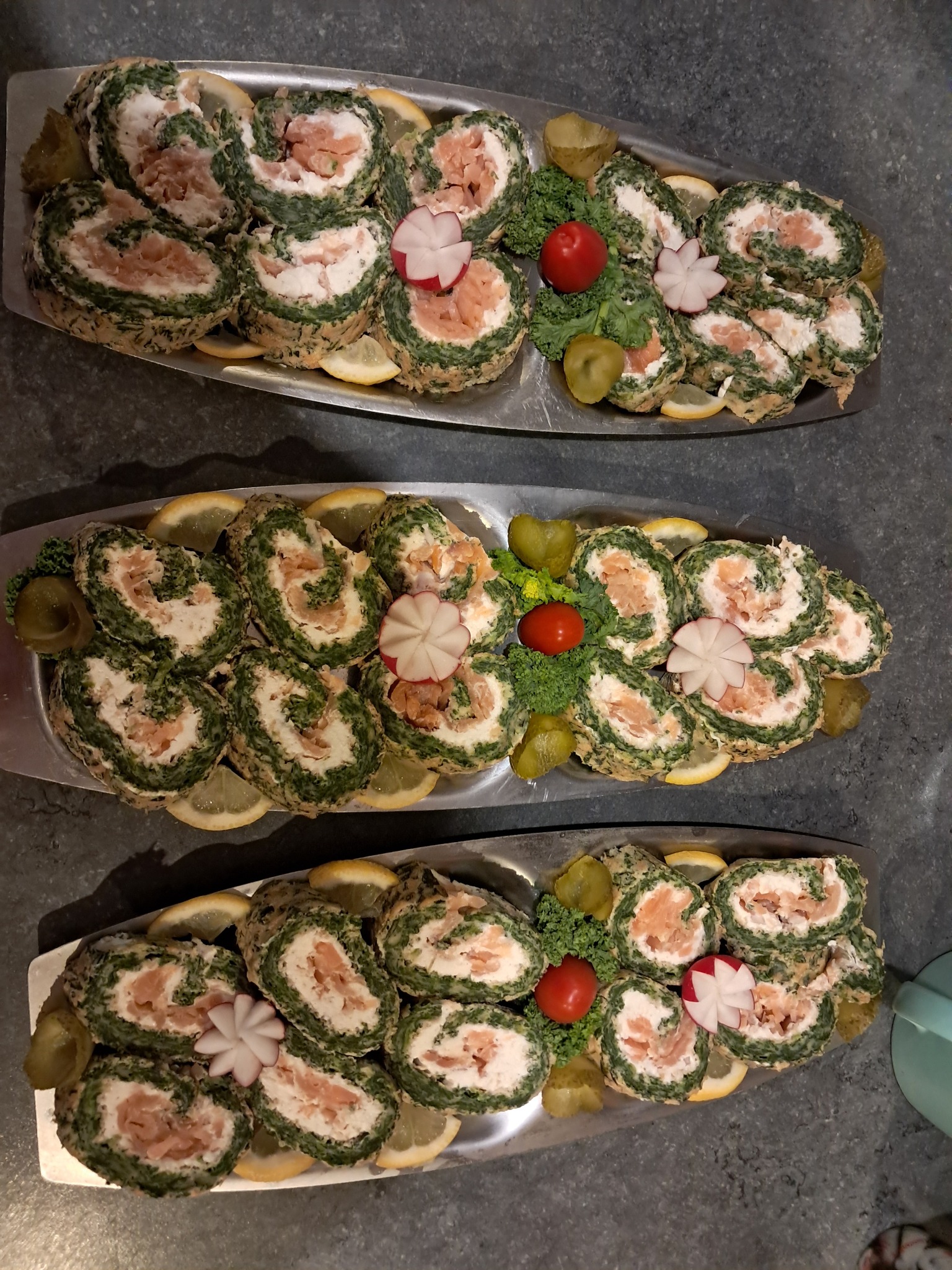Trzy srebrne tace z roladkami ze szpinakiem i łososiem, udekorowane cytryną, rzodkiewką i ogórkami konserwowymi. Widok z góry, światło naturalne.