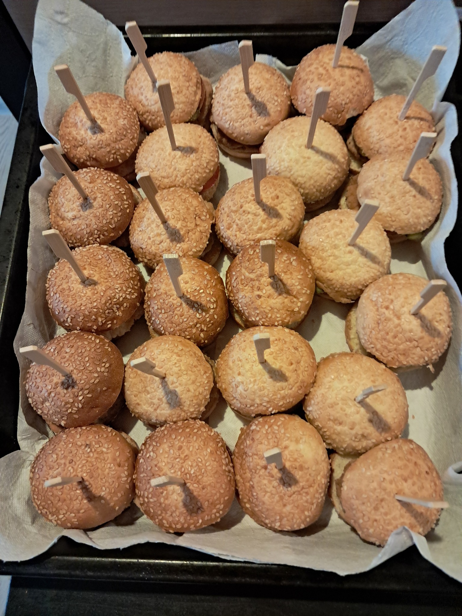 Mini burgery z sezamem na papierowej wyściółce, każdy z dekoracyjnym patyczkiem. Idealne na przyjęcia i eventy.