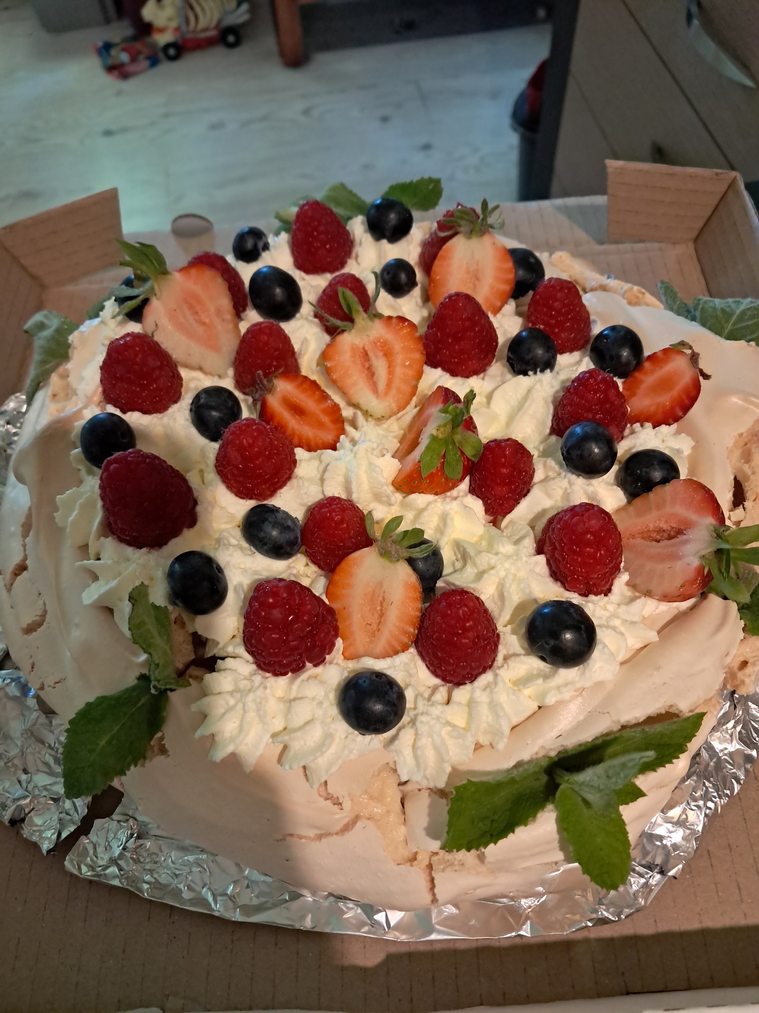 Tort Pavlova z bitą śmietaną, malinami, borówkami i truskawkami, udekorowany świeżą miętą, zapakowany w karton.