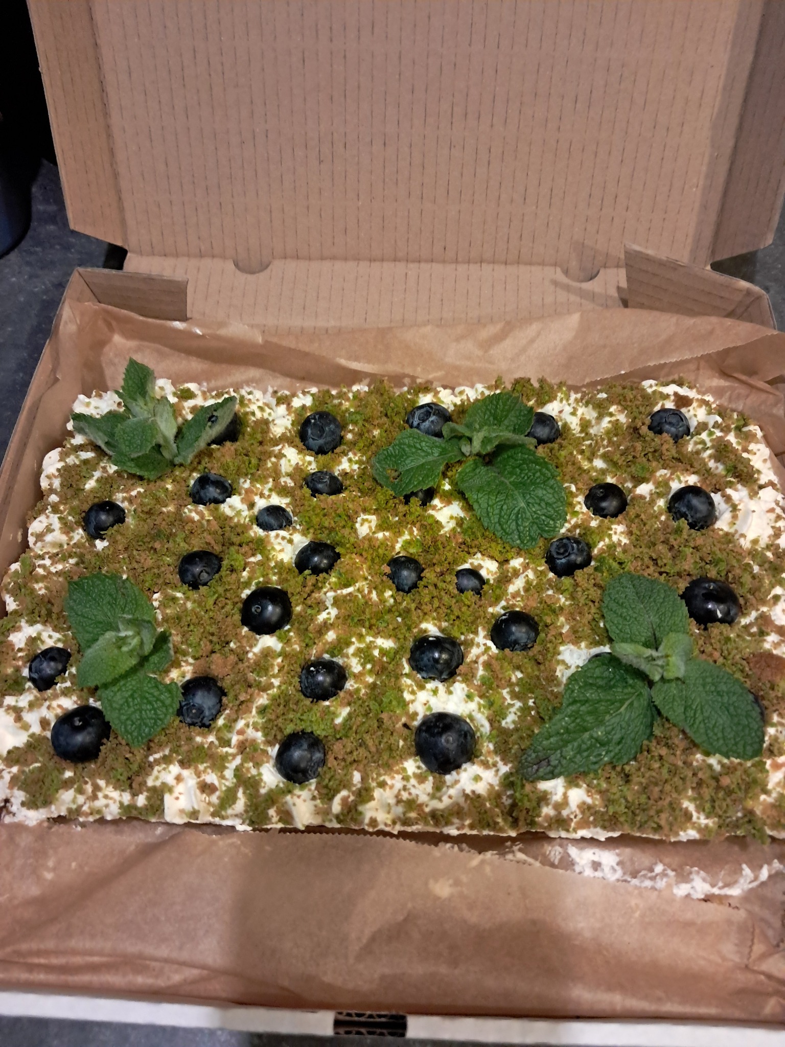 Kremowy tort z borówkami, posypką i miętą w kartonowym pudełku. Widok z góry ukazuje dekorację i świeżość składników.