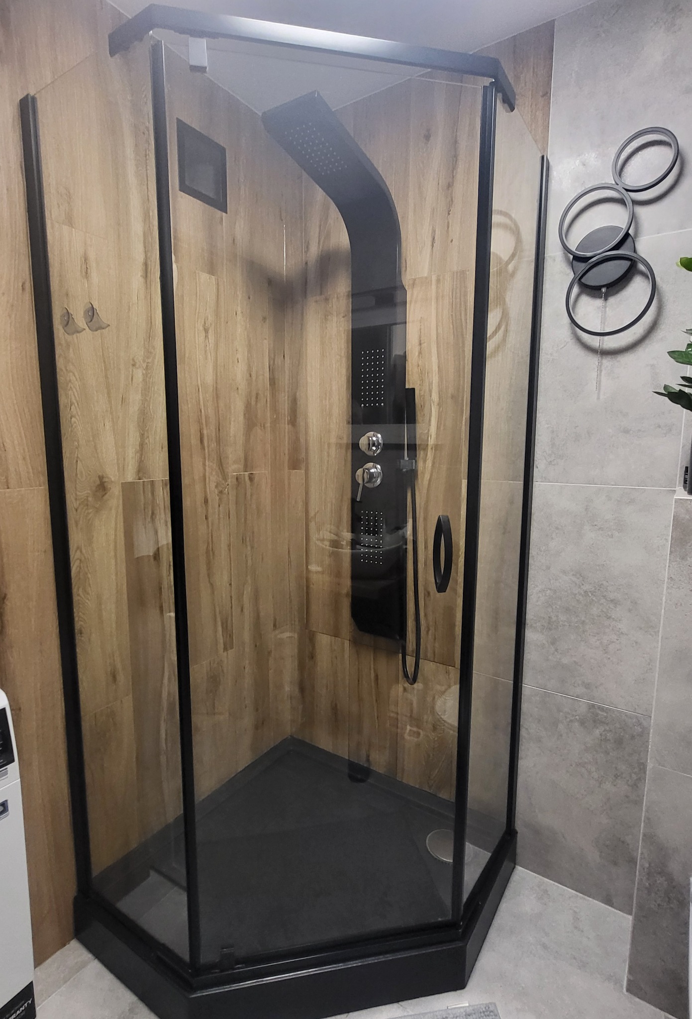 Nowoczesna kabina prysznicowa z czarną armaturą i deszczownicą, wykończona płytkami imitującymi drewno i szarym kamieniem, dekoracja ścienna w postaci metalowych kół.