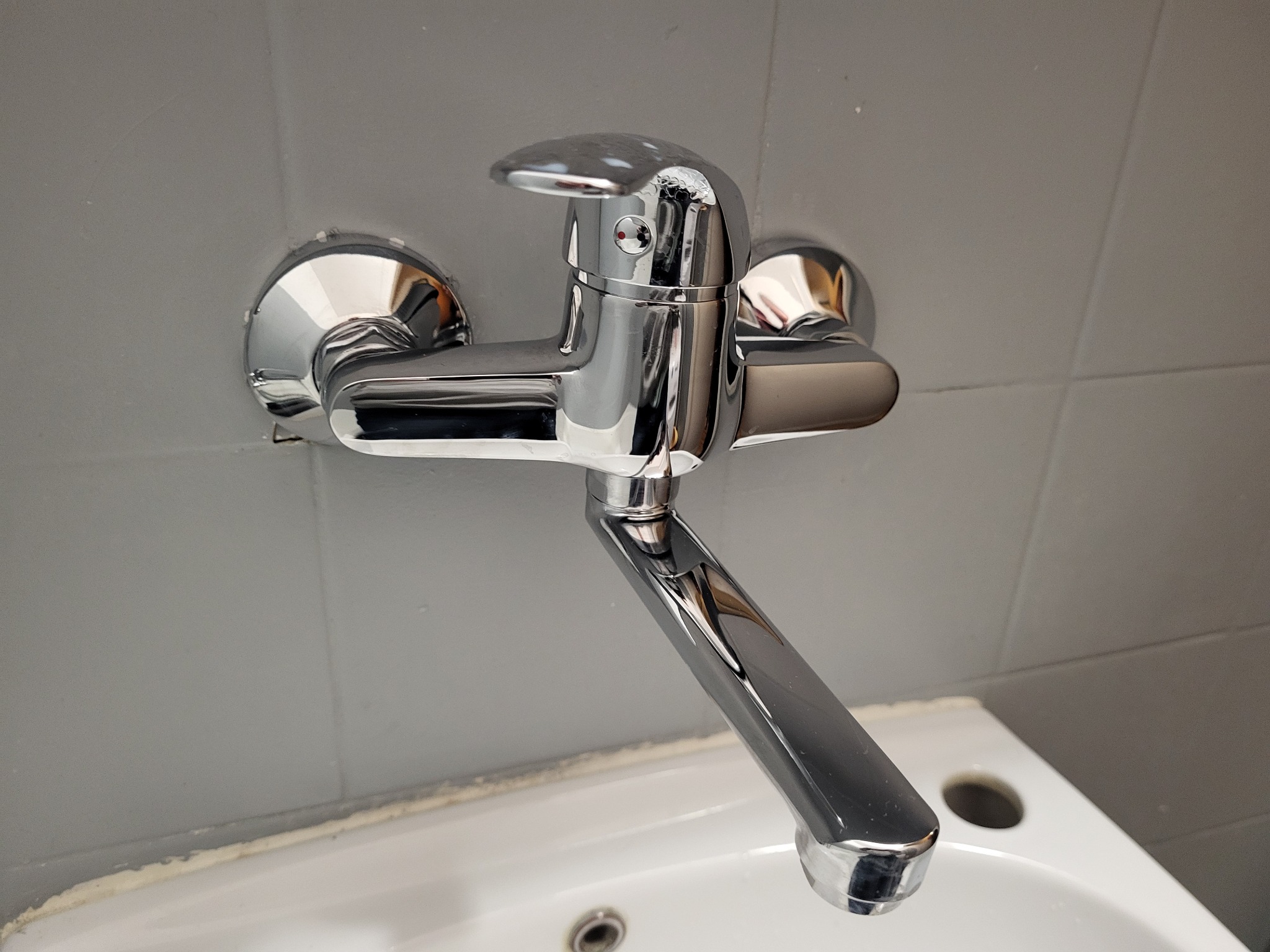 Bateria kuchenna chromowana, zamontowana na szarej ścianie nad białą umywalką z widocznymi otworami.