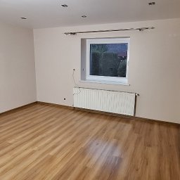 Usługi Remontowo-Budowlane GLAZUR-EFEKT Michał Sznura - Neutralnie pomalowane wnętrze pokoju z drewnianą podłogą, białym grzejnikiem pod oknem i przygotowanymi otworami na kable na ścianie.