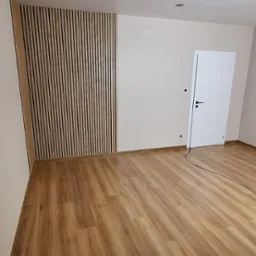 Nowoczesne wnętrze z drewnianą podłogą i dekoracyjną ścianą z pionowych lameli w odcieniach brązu i beżu, uzupełnione białą ścianą i drzwiami.