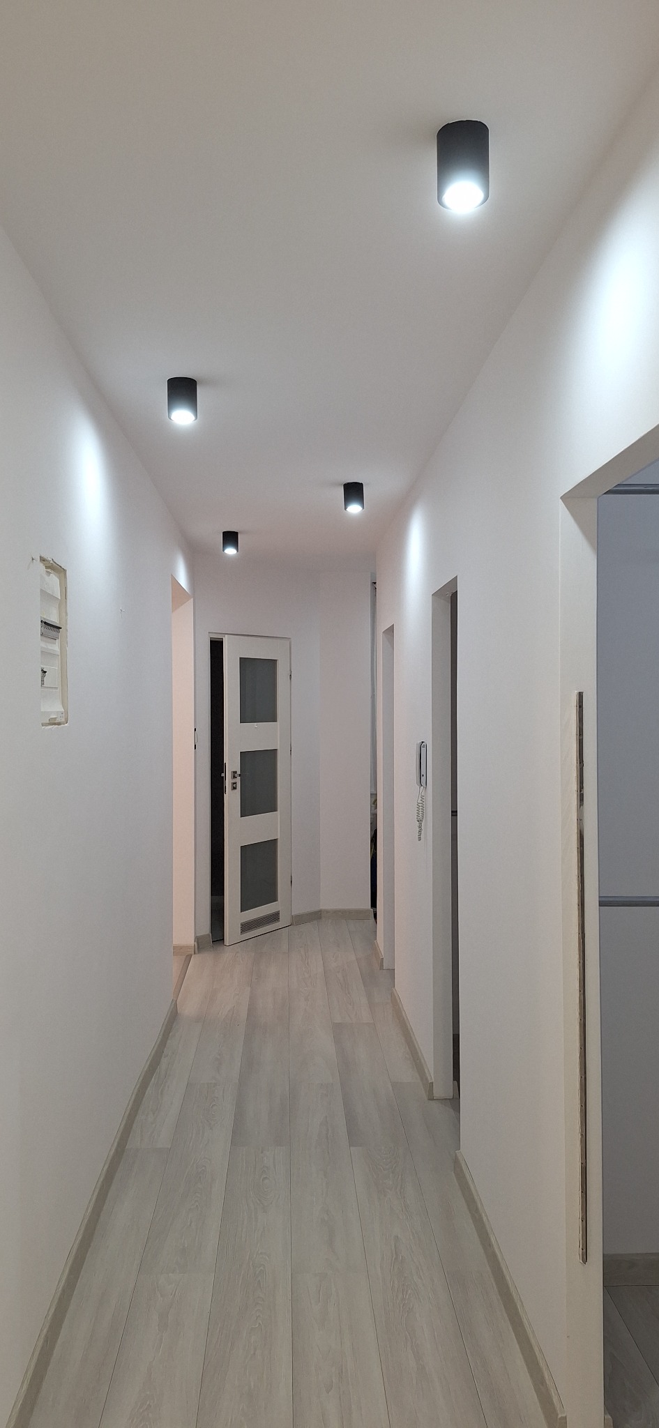 Korytarz po remoncie z parkietem w odcieniach szarości, białe ściany z czarnymi, okrągłymi lampami sufitowymi, widoczne drzwi z szybami i wystający fragment instalacji elektrycznej.