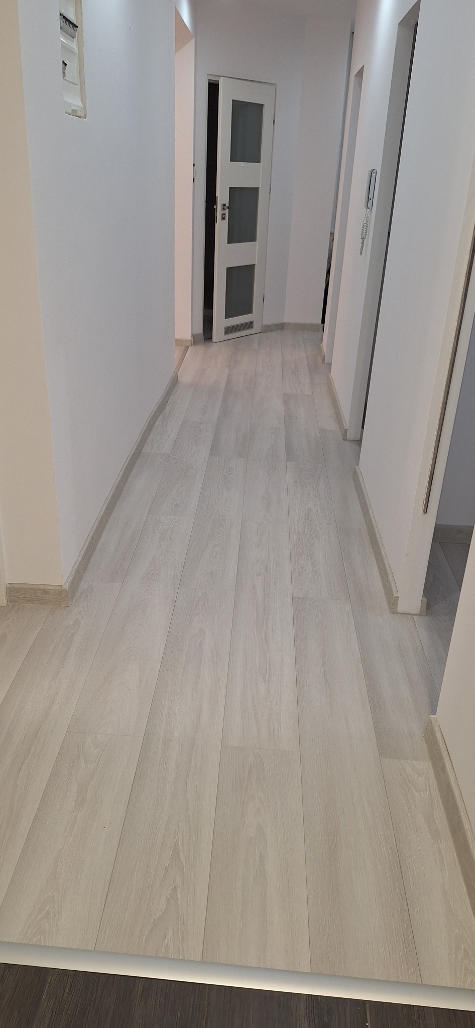 Korytarz po remoncie z panelami podłogowymi w odcieniach jasnego drewna i świeżo pomalowanymi białymi ścianami, widoczne listwy przypodłogowe i drzwi z przeszkleniami.