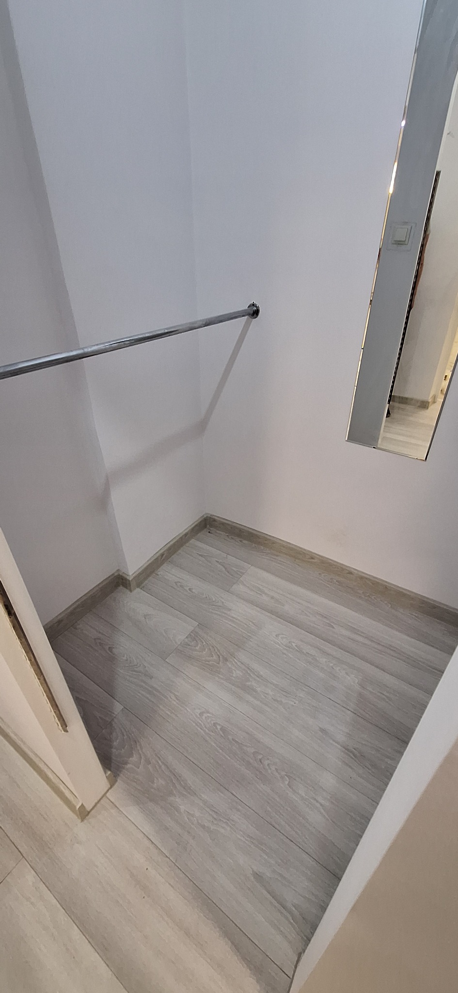 Wnęka garderoby z zamontowanym drążkiem na ubrania i lustrem na ścianie, wykończona panelami podłogowymi w odcieniach szarości.