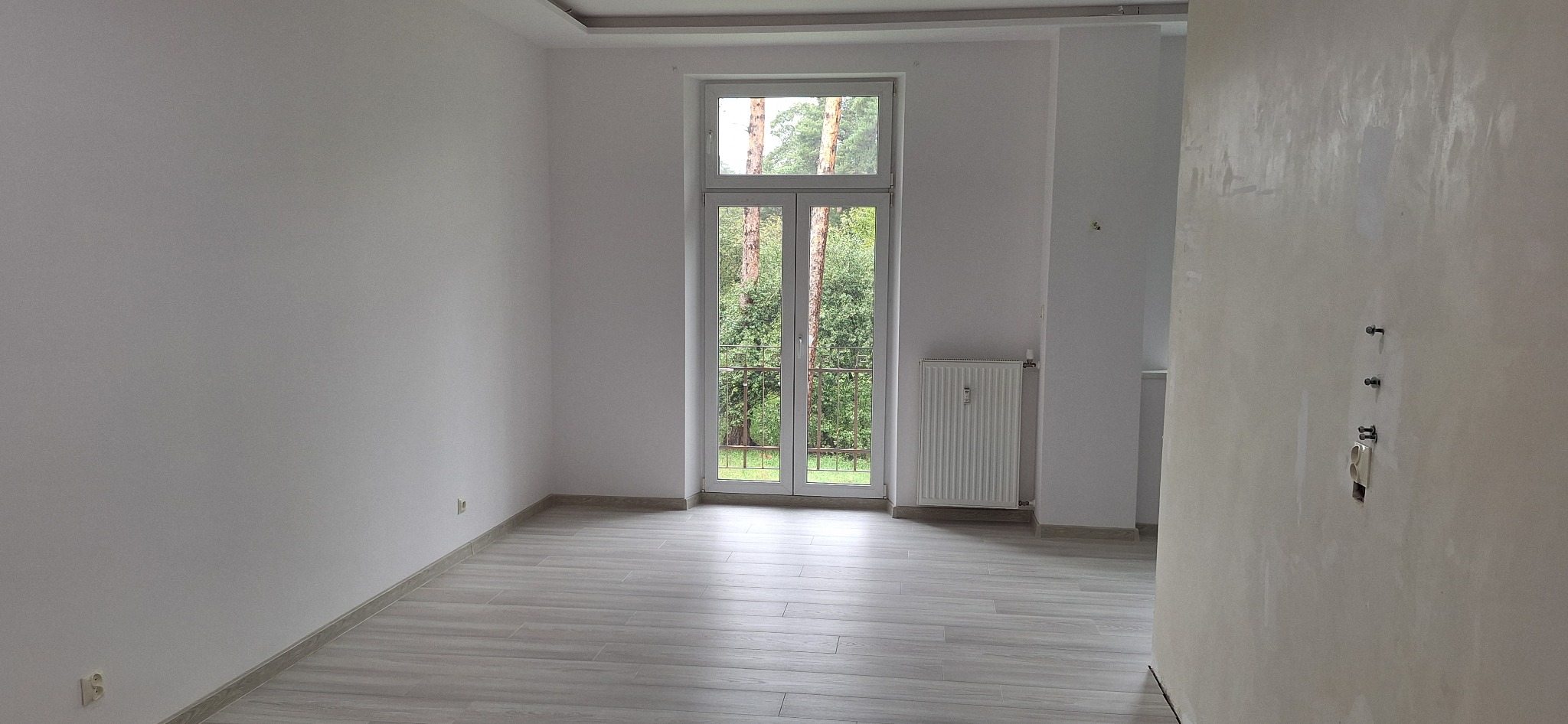 Wnętrze pokoju po remoncie, ściany pomalowane na biało, podłoga wyłożona panelami imitującymi drewno, widoczne okno balkonowe z widokiem na zieleń, grzejnik oraz fragment ściany ze śladami po...