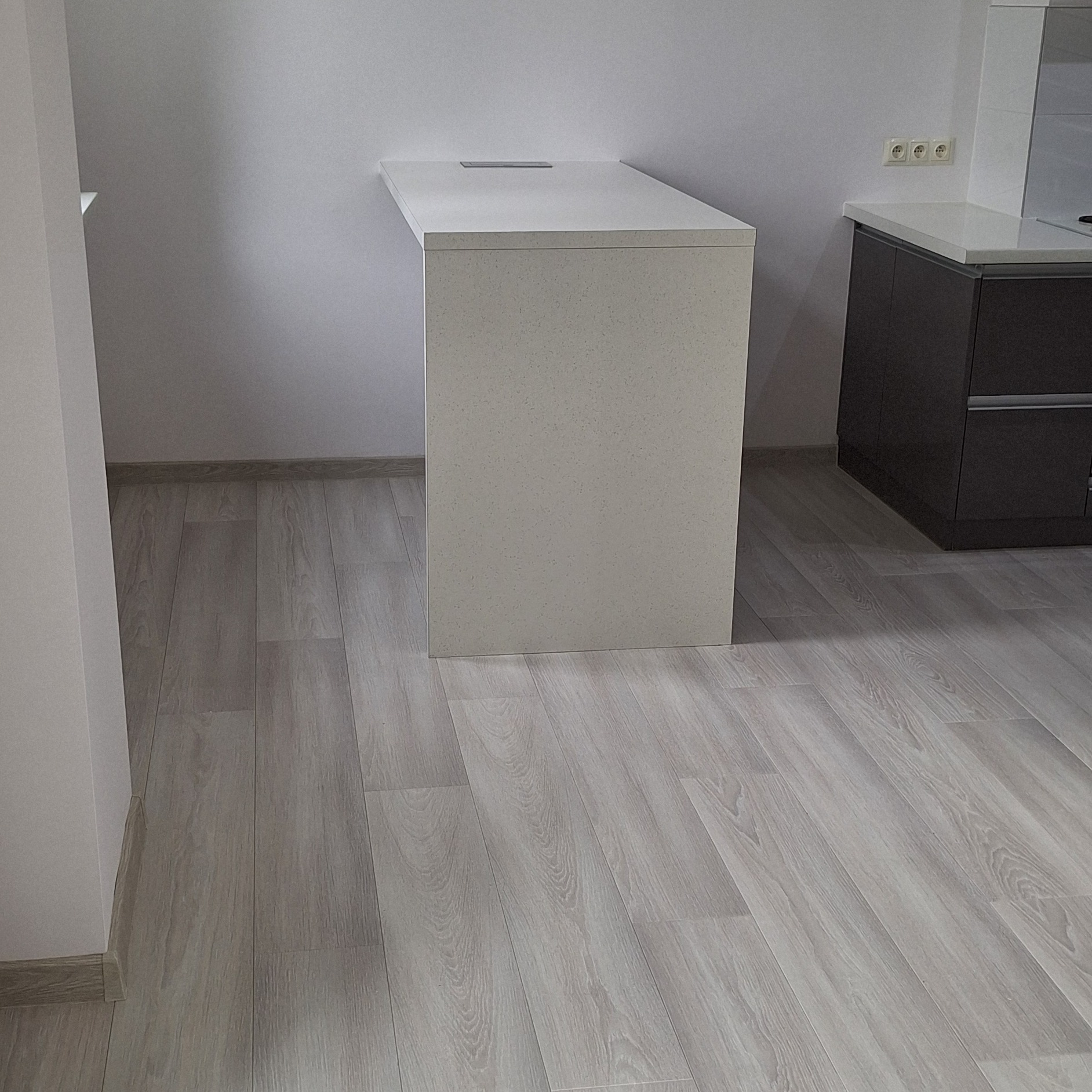 Minimalistyczna kuchnia z blatem z konglomeratu kwarcowego w odcieniach szarości, widoczne gniazdka elektryczne i podłoga z paneli imitujących drewno.