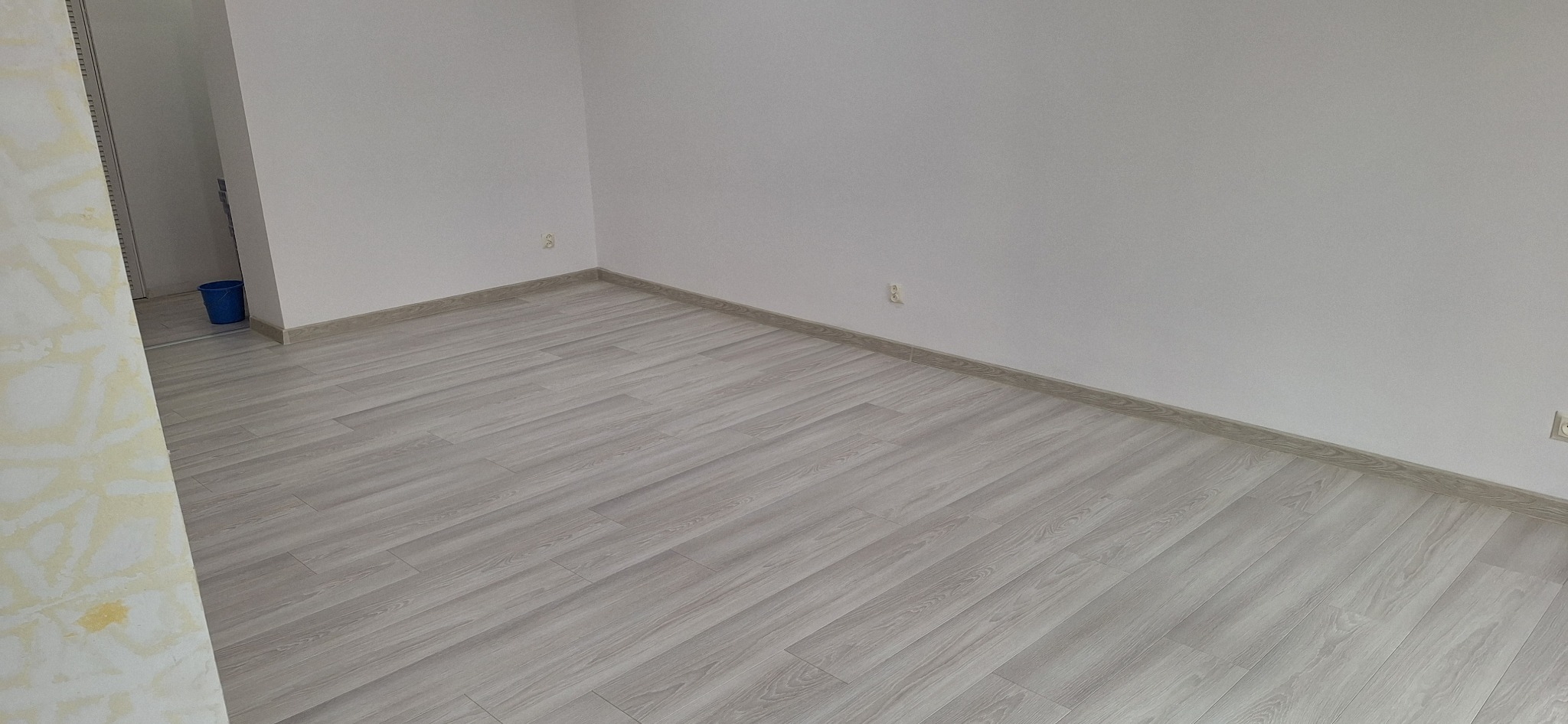Pusty pokój po remoncie, z jasnymi panelami podłogowymi imitującymi drewno, świeżo pomalowanymi białymi ścianami i widocznym niebieskim wiadrem w przejściu do innego pomieszczenia.