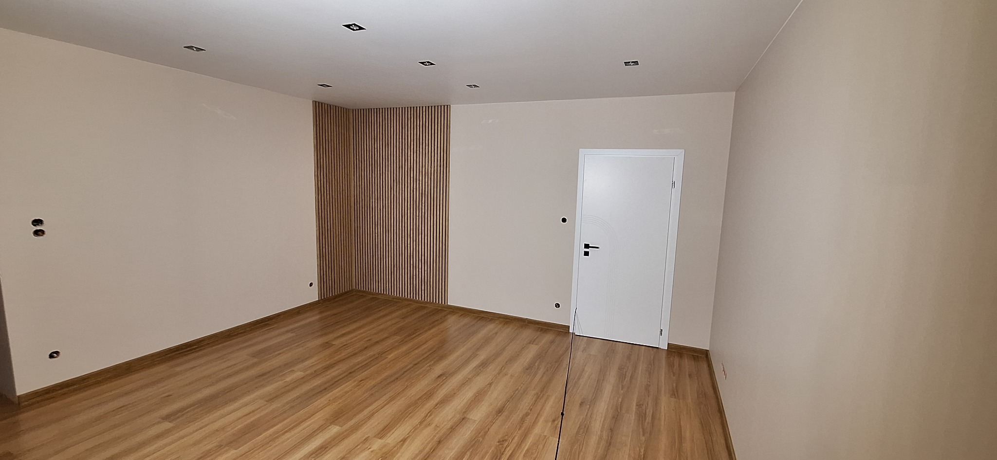 Puste pomieszczenie po remoncie: świeżo położona drewniana podłoga, beżowe ściany, białe drzwi z delikatnym wzorem, dekoracyjna ściana z drewnianych listewek oraz zamontowane oświetlenie sufitowe.