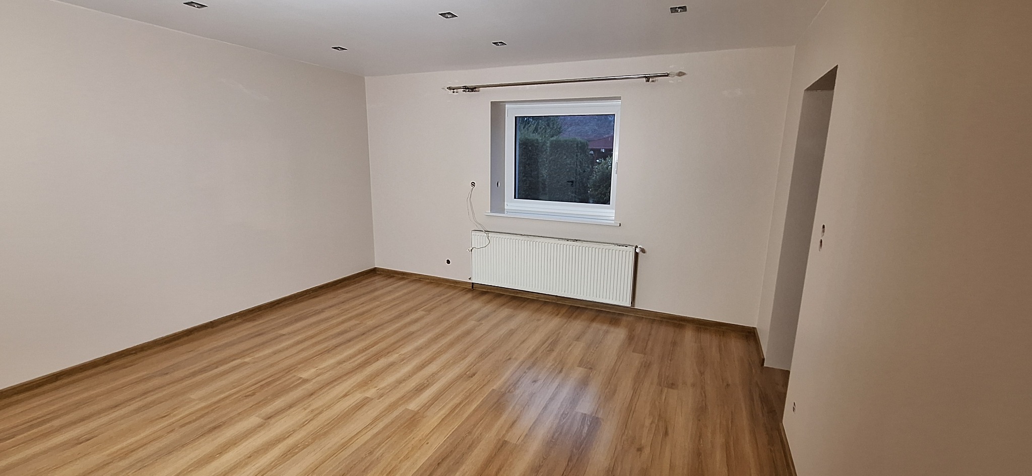 Neutralnie pomalowane wnętrze pokoju z drewnianą podłogą, białym grzejnikiem pod oknem i przygotowanymi otworami na kable na ścianie.