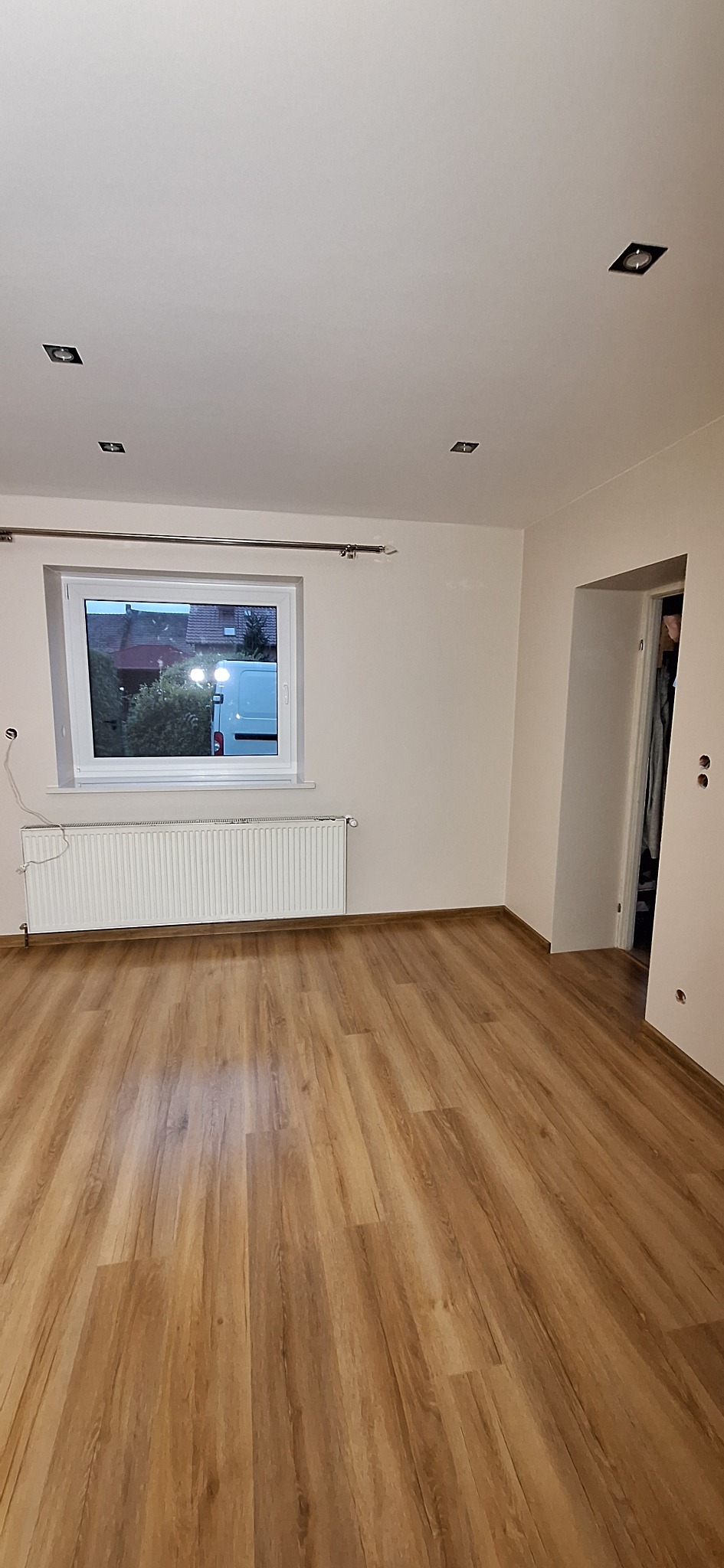 Pusty pokój po remoncie z panelami podłogowymi w odcieniach brązu, białymi ścianami, grzejnikiem pod oknem i wbudowanym oświetleniem sufitowym.