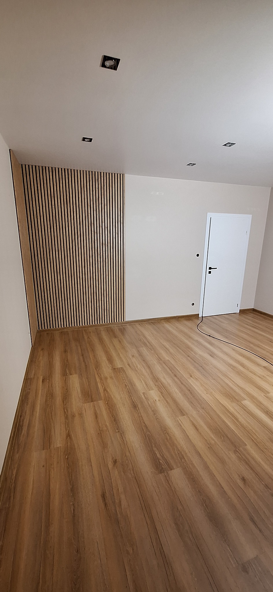 Nowoczesne wnętrze z drewnianą podłogą i dekoracyjną ścianą z pionowych lameli w odcieniach brązu i beżu, uzupełnione białą ścianą i drzwiami.