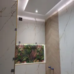 Wnętrze łazienki w trakcie wykańczania, z białymi marmurkowymi płytkami, drewnopodobną ścianą ułożoną w jodełkę, wnęką z motywem roślinnym i podwieszanym sufitem z oświetleniem LED.